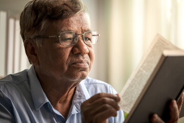 Fototapeta premium Elderly Asian man wearing glasses sitting on sofa reading book in living room at home. Close up
