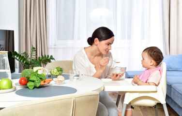 Mother feeding baby with a spoon. Spoon-feed. Happy young mother with a baby in the kitchen interior. Fresh vegetables and fruits.