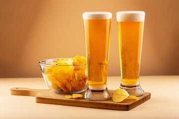 Two tall beer glasses with frothy heads and bowl of golden potato chips on wooden board against warm background. Concept of relaxation, delicious food and holiday. Two glasses beer with potato chips