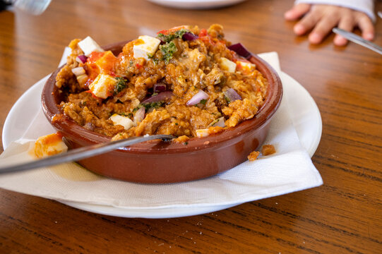 A bowl of food with a spoon in it. The bowl is on a white napkin. The food is a mixture of onion, gofio, octopus and white cheese.