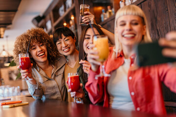 Four happy women are taking a selfie with cocktails at the bar