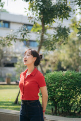 Asian woman in red shirt and jeans stands outdoors in park, looking upward with thoughtful expression, surrounded by greenery and trees