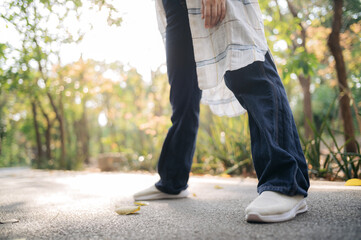 Fototapeta premium low angle view of Asian woman walking outdoors sunny day, wearing casual white shoes, blue jeans, and light plaid shirt, surrounded by greenery