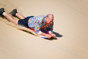 Older senior man sandboarding down sand dune on boogie board, retirement fun excitement, holiday vacation activity, healthy vital age ageing 