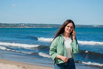 Young caucasian woman with long hair talks on phone at sunny beach, wearing a green jacket and...