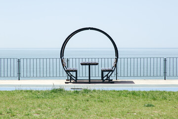 Modern round bench with a table overlooking a calm sea, placed on a promenade behind a railing, minimalistic composition with clear sky and green grass, concept of leisure