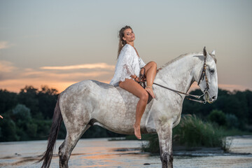 Beautiful brunette woman with long hair ,white bikini and white shirt rides  on a white horse in...