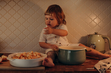 A toddler sits on a wooden countertop in a quaint kitchen, curiously touching apples while...