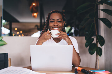 Young Black woman in white t-shirt enjoying coffee break while working remotely on laptop in cozy café, smiling and looking content during a productive freelance workday