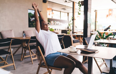 Young Black female freelancer stretching arms back in cafe after working on laptop. Relaxed and smiling, enjoying short break, casual clothes, modern remote lifestyle, coffee on table