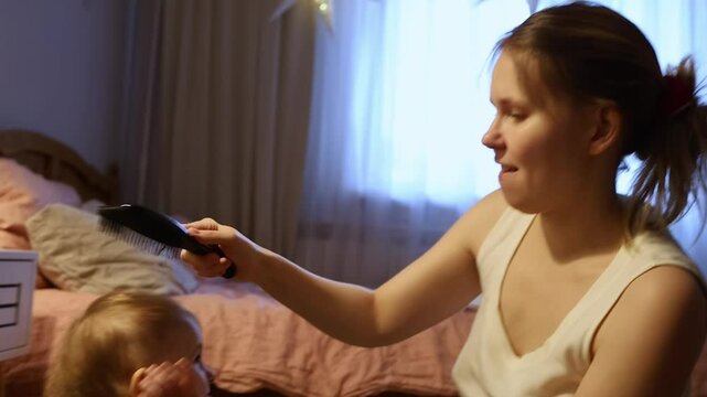 A person gently brushes the hair of a small child in a warmly lit bedroom. The child is seated, wearing a black outfit with heart patterns. Soft bedding adds to the comfort.