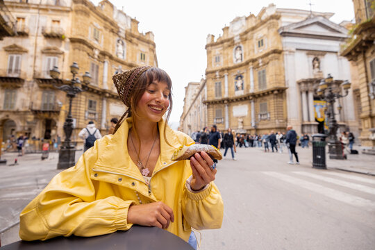 A smiling woman in a yellow jacket enjoys a traditional Sicilian cannolo while sitting at a street cafe in Palermos historic center. This image highlights authentic travel experiences, local food