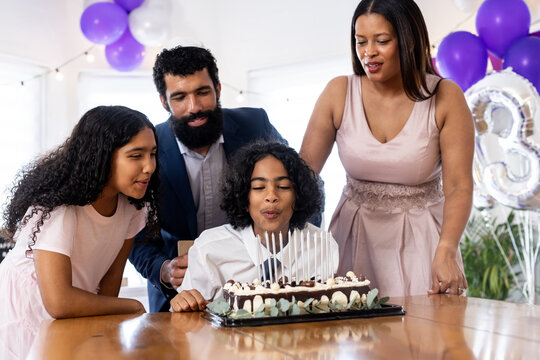 Diverse family celebrating birthday with cake and balloons, child blowing out candles, at home