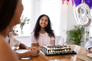 Smiling girl celebrating birthday with diverse family, sitting at table with cake, at home