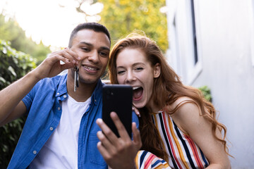 Young couple celebrating new home purchase, holding keys and taking selfie outside