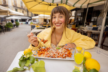 A smiling woman enjoys a traditional Pasta alla Norma at an outdoor restaurant in Sicily. The image...