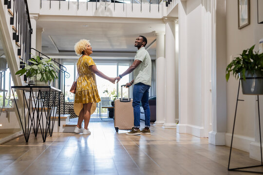 African American Couple holding hands, smiling, and entering modern home with suitcase - Powered by Adobe