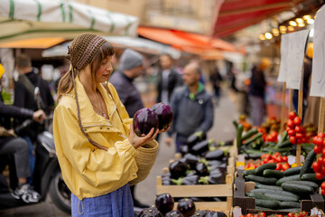 A woman in a yellow jacket selects an eggplant at a vibrant street market. This image reflects...