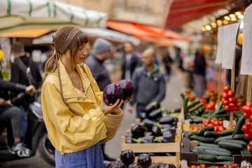 Fotobehang A woman in a yellow jacket selects an eggplant at a vibrant street market. This image reflects mindful living, healthy choices, and the charm of local food culture © rh2010