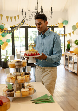 Young Indian man holding birthday cake in bright indoor party room, with cupcakes and balloons