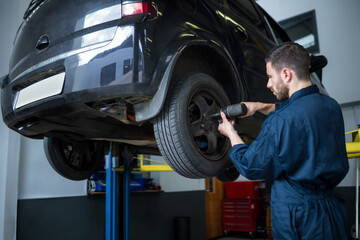 Male mechanic removing lug nuts on wheel in garage, with hydraulic lift, impact wrench, copy space