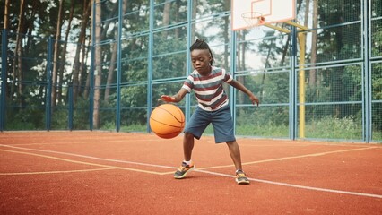 Close up of little boy bouncing ball outside in city park on basketball court. African American kid practicing playing basketball outdoor. Sport and recreation. Leisure concept. Professional sport