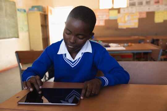 Working African American schoolboy tapping tablet at classroom desk, with chalkboard and posters