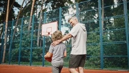 Rear of mature father and little son with basketball ball walking away from playground court. Caucasian man walks with teen son after playing basketball. Back view. Sport concept. Family activity