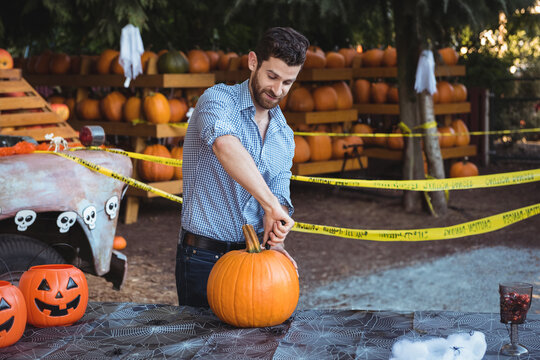 Man carving pumpkin at outdoor market stall, with patterned tablecloth, knife and jack-o'-lanterns