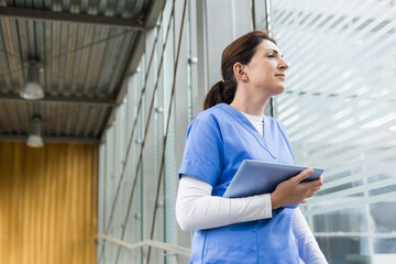Standing female nurse reviewing patient data in medical corridor, with tablet computer, copy space