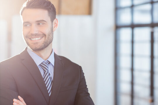 Smiling man standing with folded arms in modern office, with dark suit jacket and striped tie