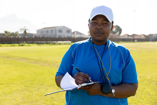 African American woman writing on clipboard on sports field, with whistle and stopwatch, copy space