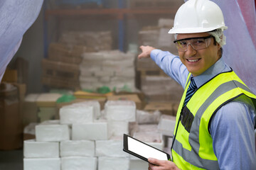 Pointing man inspecting stock in warehouse wearing safety vest, with tablet and boxes, copy space