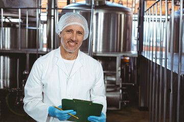 Inspecting male technician examining fermentation tanks in brewery, with green clipboard and pencil