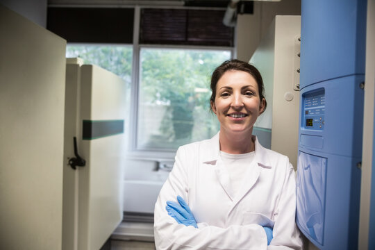 Smiling woman wearing lab coat and gloves in laboratory, highlighting freezer and control panel