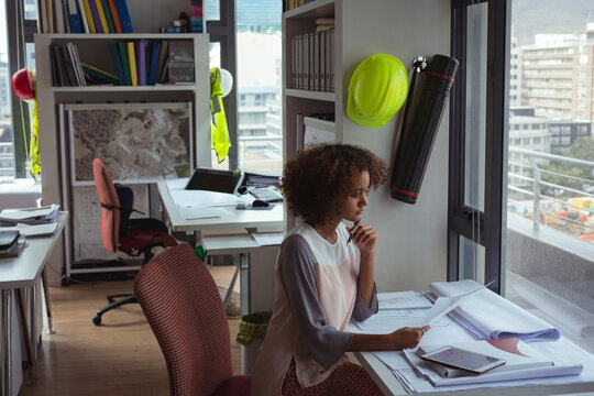 Architect reviewing blueprints in studio with laptop, tablet and safety helmet near window