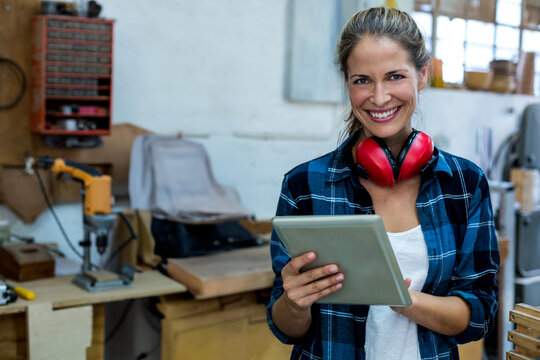 Woman wearing plaid shirt and red earmuffs holding tablet in wood shop, copy space