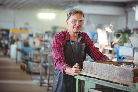 Leaning senior man wearing safety goggles at industrial workshop, with wooden crate of metal parts