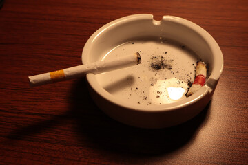A cigarette rests in a white ashtray, surrounded by ash and the remnants of a finished smoke. The dim lighting adds a moody, contemplative atmosphere.