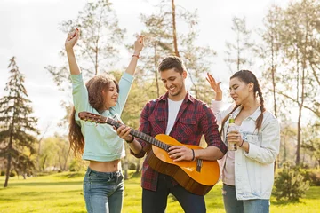 Zelfklevend Fotobehang Three friends enjoy a sunny outdoor day in the park, playing guitar, singing, and dancing, showcasing joy and togetherness in a natural and relaxed setting. © kegfire