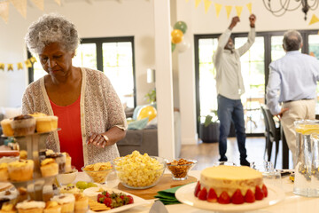 Elderly woman arranging cupcakes on table while guests celebrating in background