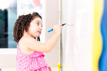 Cute toddler girl drawing on whiteboard in kindergarten classroom