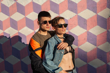 A stylish couple wearing sunglasses poses confidently against a vibrant geometric mural outdoors,...