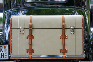 A vintage beige travel trunk with leather straps mounted on the back of a black classic car. The...