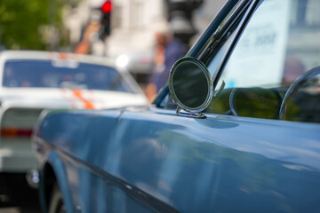 Side view of a classic blue car with the round chrome side mirror in focus. Blurred street and car dealership in the background.