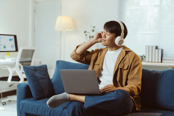 Young Asian man enjoying music with headphones and laptop while working remotely from his comfortable home office