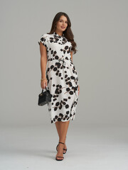 Elegant woman in a knee-length white dress with black floral patterns, holding a small black...