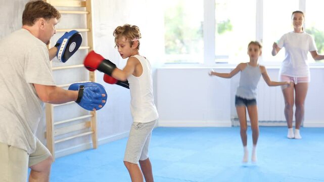Boy and dad are boxing in gym, father helps child to work out force of blow. Modern positive family chooses active hobby
