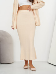 Woman in cream-colored skirt and cardigan with white top. She stands near a gray modern chair and...