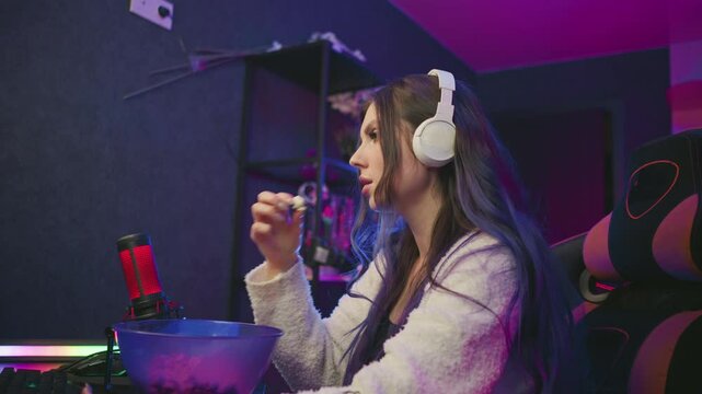 Young woman wearing headphones eats popcorn from bowl while working at computer desk in vibrant studio setup with bright LED lighting and microphone